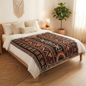 May include: A woven throw blanket featuring a repeating geometric pattern in shades of black, brown, and tan. The blanket has a fringed edge and is draped over a white bedspread. The design includes horizontal bands of triangles, lines, and arches.