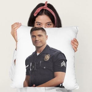 May include: White square pillow featuring a man in a police uniform. The man is wearing a dark blue short-sleeved shirt with a badge and stripes on the sleeve. The pillow is held by a person.