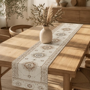 May include: A long, rectangular table runner featuring a cream-colored fabric base with a repeating pattern of brown and dark gray geometric medallions and diamond-shaped borders. The runner is centered on a wooden table with a smooth finish.