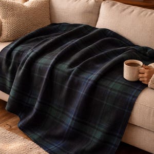 May include: A dark blue and green plaid blanket draped over a beige sofa. The blanket has a classic tartan pattern. A beige pillow rests on the sofa, and a mug of coffee sits on the blanket. The scene is lit by a warm lamp.