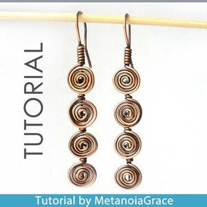 May include: A pair of copper wire earrings with four coiled circles on each earring. The earrings are hanging from a wooden dowel.