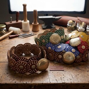 May include: Two belts made of circular leather pieces in brown, green, blue, red, and yellow, each featuring small metal studs and a large, round, embossed brass buckle. A small rectangular leather tag reads MOROCCAN ARTISAN in the center.