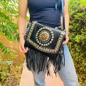 Könnte beinhalten: Schwarze Leder-Umhängetasche mit silbernen Nieten und einem großen zentralen Schmuckelement. Die Tasche hat einen Klappverschluss und lange schwarze Fransen am unteren Rand. Ein stilvolles Accessoire für den täglichen Gebrauch.