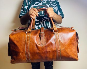 May include: A large, brown leather duffel bag with a buckle closure and two top handles. The bag has a rectangular shape with visible stitching and a textured surface. The bag is held by a person wearing a green and white patterned shirt.