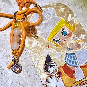 May include: An orange keychain with a cartoon illustration. The keychain has a lobster clasp, a beaded charm, and a pendant with a cartoon girl and a cat. The illustration is in shades of yellow, orange, white, and blue.