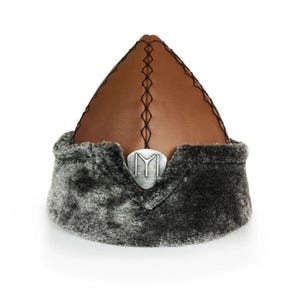 May include: A brown leather hat with a pointed crown and grey fur trim. The hat has a silver emblem at the front and black stitching along the top seam. This traditional hat is a distinctive accessory.