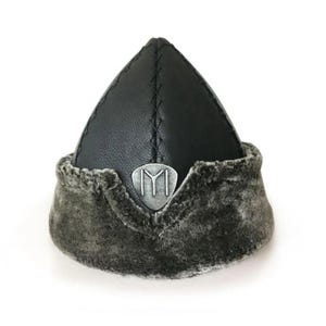 May include: A black leather hat with a pointed crown and a faux fur trim. A silver emblem with a stylized letter "I" is centered on the front. The hat is designed in a traditional style.