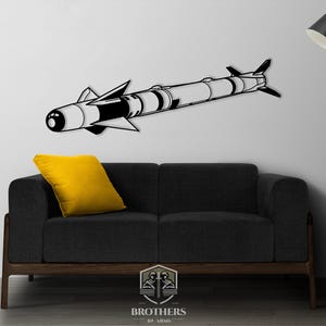 May include: A black and white metal wall art piece depicting a missile. The artwork is mounted on a white wall above a dark gray sofa with a yellow pillow. A black floor lamp with a gold accent is to the right.