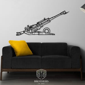 Puede incluir: Arte de pared de metal negro que representa un cañón en posición de disparo. El cañón está montado en un carro con ruedas y tiene un cañón largo. El arte está montado en una pared blanca sobre un sofá negro con un cojín amarillo.
