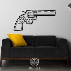 May include: Black metal wall art of a revolver pistol with a white outline. The art is mounted on a white wall above a black couch with a yellow throw pillow. The art is made by Brothers In Arms.