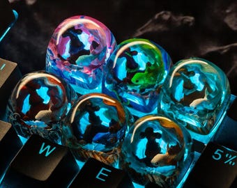 May include: Six colorful resin keycaps with a blue, green, pink, and orange background. Each keycap features a unique design of a fish swimming in a blue, green, or orange ocean. The keycaps are arranged on a black keyboard with the keys 'W', 'E', and 'R' visible.