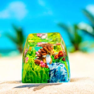 May include: A clear, rectangular resin keycap featuring a miniature landscape with a small, brown and tan creature. The scene includes green grass, pink flowers, and a blue waterfall, set against a blurred beach and sky background.
