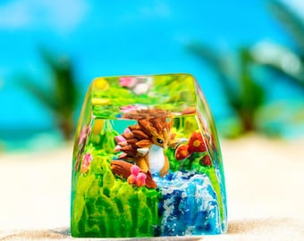 May include: A clear, rectangular resin keycap featuring a miniature landscape with a small, brown and tan creature. The scene includes green grass, pink flowers, and a blue waterfall, set against a blurred beach and sky background.