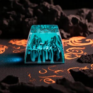 May include: A teal-coloured keycap with a mountain range design. The square-shaped keycap has a detailed, layered mountain scene within its clear resin body. The background is dark with orange swirls. The keycap is approximately 2.5 cm square.