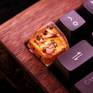 May include: A clear resin keycap with a honeycomb design and two bees inside. The keycap is on a wooden keyboard.