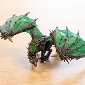 May include: A miniature dragon figurine featuring a black body with sharp, textured spikes along its back and head. The dragon has large, outstretched wings with a vibrant green membrane and dark, veined detailing, finished with pointed gold claws.