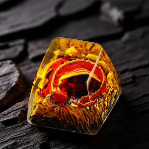 May include: A clear, geometric resin art piece with a vibrant, layered design. The interior features a red and yellow floral or abstract form, encased in transparent resin. The piece is set against a dark, textured background.
