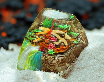 May include: A clear resin keycap featuring a miniature landscape with a dragonfly, waterfall, and colorful plants. The keycap has a textured, rocky base and is set against a backdrop of sand.