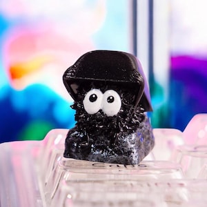 Op de afbeelding: Een zwarte en witte Totoro-toetsdop met een zwarte hoed en grote witte ogen. De toetsdop zit op een toetsenbord met het woord "Caps" zichtbaar.