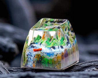 May include: A clear, rectangular resin keycap with a miniature landscape scene inside. The scene features green, blue, and orange painted mountains, a river, and a small red boat. The keycap is set against a dark, textured background.