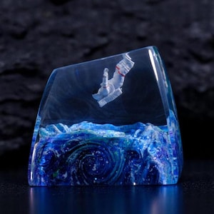 May include: A clear resin sculpture depicting an astronaut floating above a swirling blue ocean. The astronaut is white with red detailing. The base of the sculpture is a deep blue, with white and blue swirls.