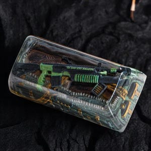 May include: A clear resin block with a green and black toy gun inside. The gun is surrounded by brown and green fabric and a magazine of bullets.