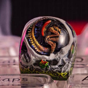 May include: A clear resin keycap with a detailed, gold-colored alien creature inside. The creature is curled up in a fetal position, with its tail wrapped around its body. The keycap is set on a keyboard with the word "caps" visible.