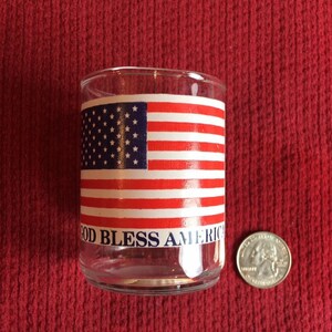 May include: A clear glass tumbler with a patriotic design featuring the American flag and the words "GOD BLESS AMERICA." A quarter is next to the glass for size comparison. The glass sits on a red surface.