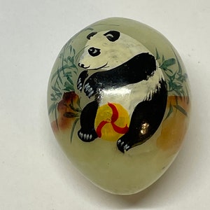 May include: Oval-shaped decorative item featuring a hand-painted panda bear. The panda, with black and white fur, is holding a yellow ball with red accents. The background has green bamboo and floral elements.