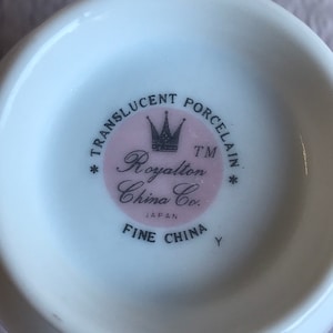 May include: A close-up of the bottom of a white porcelain teacup. The center features a pink circle with the words "TRANSLUCENT PORCELAIN" and "Royalton China Co. JAPAN FINE CHINA" in black text. A crown graphic is in the center.