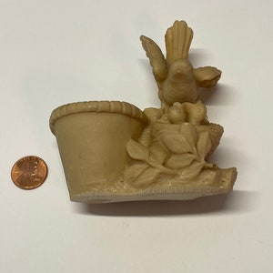 Peut inclure: Figurine d&eacute;corative beige repr&eacute;sentant un oiseau au-dessus d'un petit pot. L'oiseau a les ailes d&eacute;ploy&eacute;es et est entour&eacute; de feuilles et de fruits sculpt&eacute;s. Le pot a un bord textur&eacute;. Une pi&egrave;ce de monnaie est &agrave; c&ocirc;t&eacute; de la figurine pour l'&eacute;chelle.