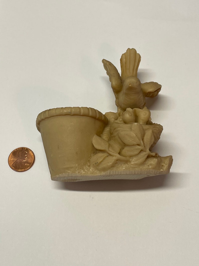 Peut inclure: Figurine d&eacute;corative beige repr&eacute;sentant un oiseau au-dessus d'un petit pot. L'oiseau a les ailes d&eacute;ploy&eacute;es et est entour&eacute; de feuilles et de fruits sculpt&eacute;s. Le pot a un bord textur&eacute;. Une pi&egrave;ce de monnaie est &agrave; c&ocirc;t&eacute; de la figurine pour l'&eacute;chelle.