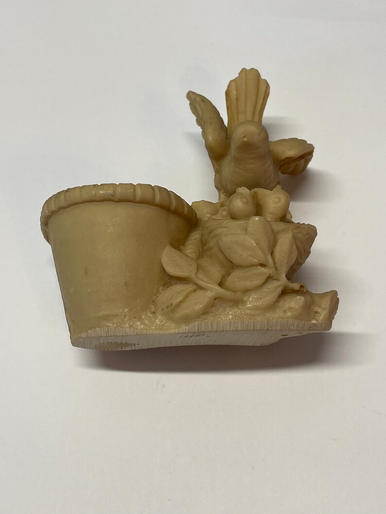 Peut inclure: Figurine beige repr&eacute;sentant un oiseau sur un nid avec un petit panier. L'oiseau a les ailes d&eacute;ploy&eacute;es et est entour&eacute; de feuilles et de petits fruits. Le panier est sur le c&ocirc;t&eacute; gauche.