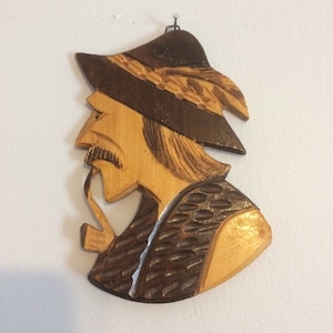 May include: Wooden wall hanging depicting a man in profile, wearing a hat and smoking a pipe. The carving features a dark brown hat and vest, with the face and hair in a lighter wood tone. The piece is designed to be hung on a wall.