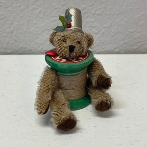 May include: A whimsical teddy bear figurine with a silver thimble hat and a green and red spool body. The bear has brown fur and dark brown paws. A red button and holly sprig add festive detail. A unique holiday decoration.