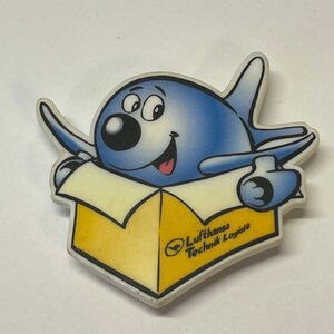 May include: A blue cartoon airplane character with a smiling face and outstretched arms, emerging from a yellow box. The box has the text "Lufthansa Technik Logistik" and a logo.