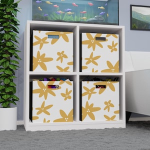 Peut inclure: Unité de rangement blanche avec quatre compartiments, chacun contenant un bac de rangement à motifs. Les bacs présentent un fond blanc avec un motif floral doré répété. L'unité est placée sur un tapis gris.