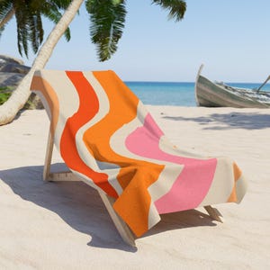 Peut inclure: Une serviette de plage avec un motif rétro ondulé orange, rose et crème, drapée sur une chaise longue en bois. La scène comprend une plage de sable, l'océan et un bateau en arrière-plan.