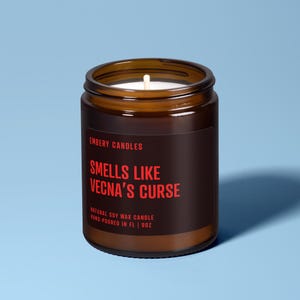 May include: A brown glass jar candle with a black label featuring red text that reads EMBERY CANDLES, SMELLS LIKE VECNA'S CURSE, NATURAL SOY WAX CANDLE, HAND-POURED IN FL | 9OZ. The candle has a single white cotton wick centered in the white wax.