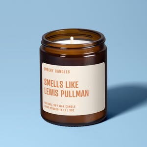 May include: A brown glass jar candle with a white label. The label reads "EMBERY CANDLES SMELLS LIKE LEWIS PULLMAN NATURAL SOY WAX CANDLE HAND-POURED IN FL | 9OZ". The candle has a white wick and is set against a light blue background.