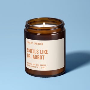 Könnte beinhalten: Eine braune Glas-Kerze mit weißem Etikett. Auf dem Etikett steht "EMBERY CANDLES SMELLS LIKE DR. ABBOT NATURAL SOY WAX CANDLE HAND-POURED IN FL | 9OZ". Die Kerze steht vor blauem Hintergrund.