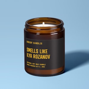 May include: A brown glass jar candle with a black label. The label reads "EMBERY CANDLES SMELLS LIKE ILYA ROZANOV NATURAL SOY WAX CANDLE HAND-POURED IN FL | 9OZ". The candle has a white wick and a blue background.