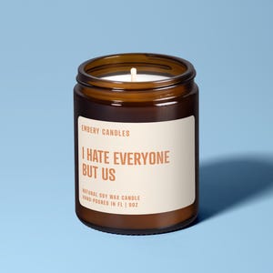 May include: A brown glass jar candle with a white label featuring the text EMBERY CANDLES, I HATE EVERYONE BUT US, NATURAL SOY WAX CANDLE, HAND-POURED IN FL | 9OZ. The candle has a single white wick centred in the white wax inside the glass container.