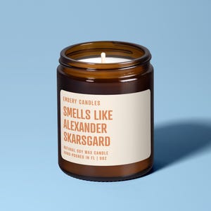 May include: A brown glass jar candle with a white label. The label reads "EMBERY CANDLES SMELLS LIKE ALEXANDER SKARSGARD NATURAL SOY WAX CANDLE HAND-POURED IN FL | 9OZ". The candle has a white wick and is set against a light blue background.