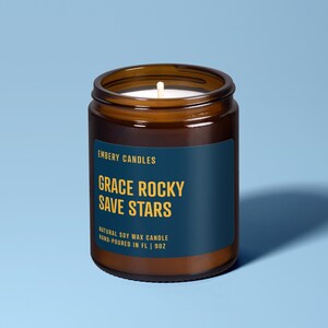 May include: A brown glass jar candle with a dark blue label featuring the text EMBERY CANDLES, GRACE ROCKY SAVE STARS, and NATURAL SOY WAX CANDLE HAND-POURED IN FL | 9OZ. The candle has a white wick centered in the wax inside the glass container.