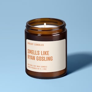 May include: A brown glass jar candle with a white label featuring the text EMBERY CANDLES, SMELLS LIKE RYAN GOSLING, NATURAL SOY WAX CANDLE, HAND-POURED IN FL | 9OZ. The candle has a single white wick centered in the white wax inside the jar.