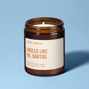 Könnte beinhalten: Eine braune Glas-Kerze mit einem weißen Etikett, das den Text EMBERY CANDLES, SMELLS LIKE DR. SANTOS, NATURAL SOY WAX CANDLE, HAND-POURED IN FL | 9OZ trägt. Die Kerze hat einen einzelnen weißen Docht, der mittig im weißen Sojawachs sitzt.