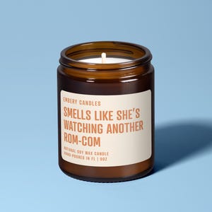 Pode incluir: Uma vela em pote de vidro castanho com um rótulo branco. O rótulo diz "SMELLS LIKE SHE'S WATCHING ANOTHER ROM-COM". A vela contém cera branca e um pavio. Em um fundo azul claro.