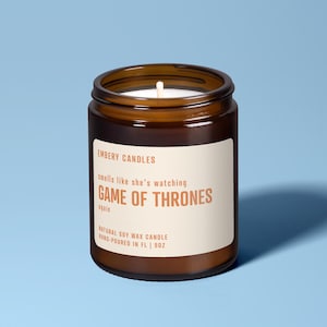 Może przedstawiać: Brązowa świeca w szklanym słoiku z białą etykietą. Na etykiecie napisano "EMBERY CANDLES" i "smells like she's watching GAME OF THRONES again." Świeca jest naturalną świecą sojową, ręcznie wylaną w FL i waży 255 gramów.