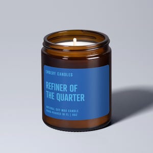 May include: A brown glass candle jar with a blue label that reads "EMBERY CANDLES REFINER OF THE QUARTER NATURAL SOY WAX CANDLE HAND-POURED IN FL | 9OZ".